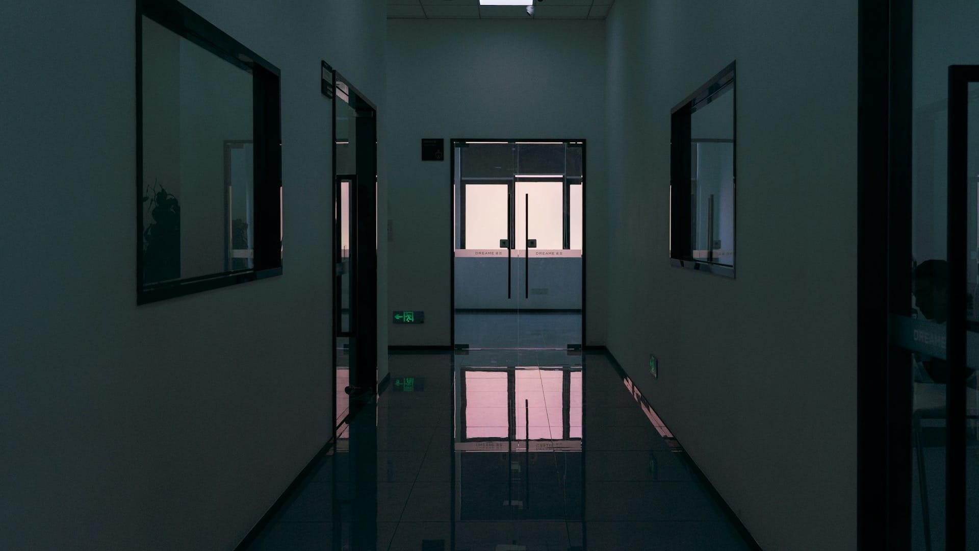 A dim office corridor ending in glass double doors, with a lit room visible through them