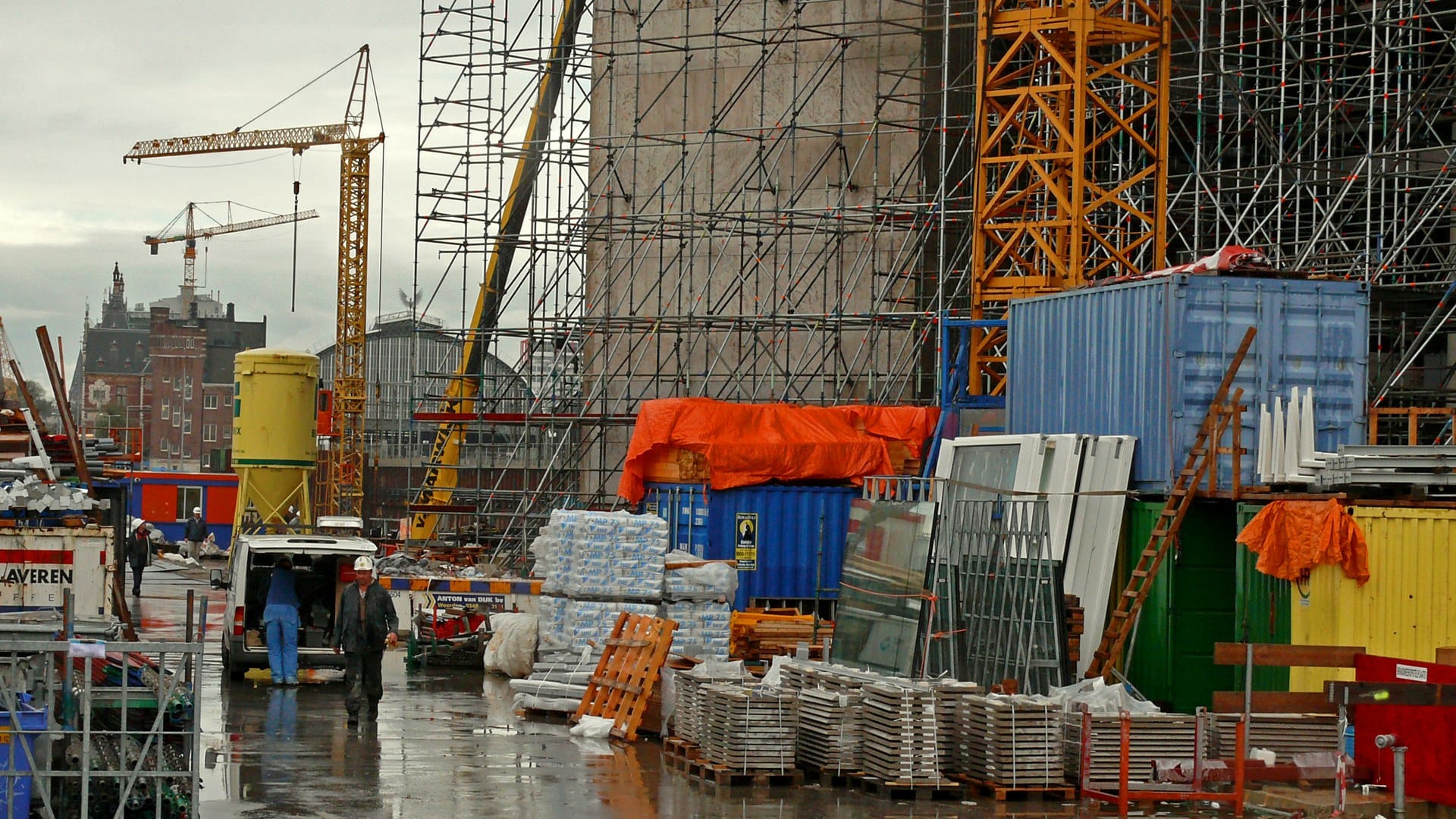 A building site under scaffolding, with cranes overhead and materials stacked on the wet ground below.