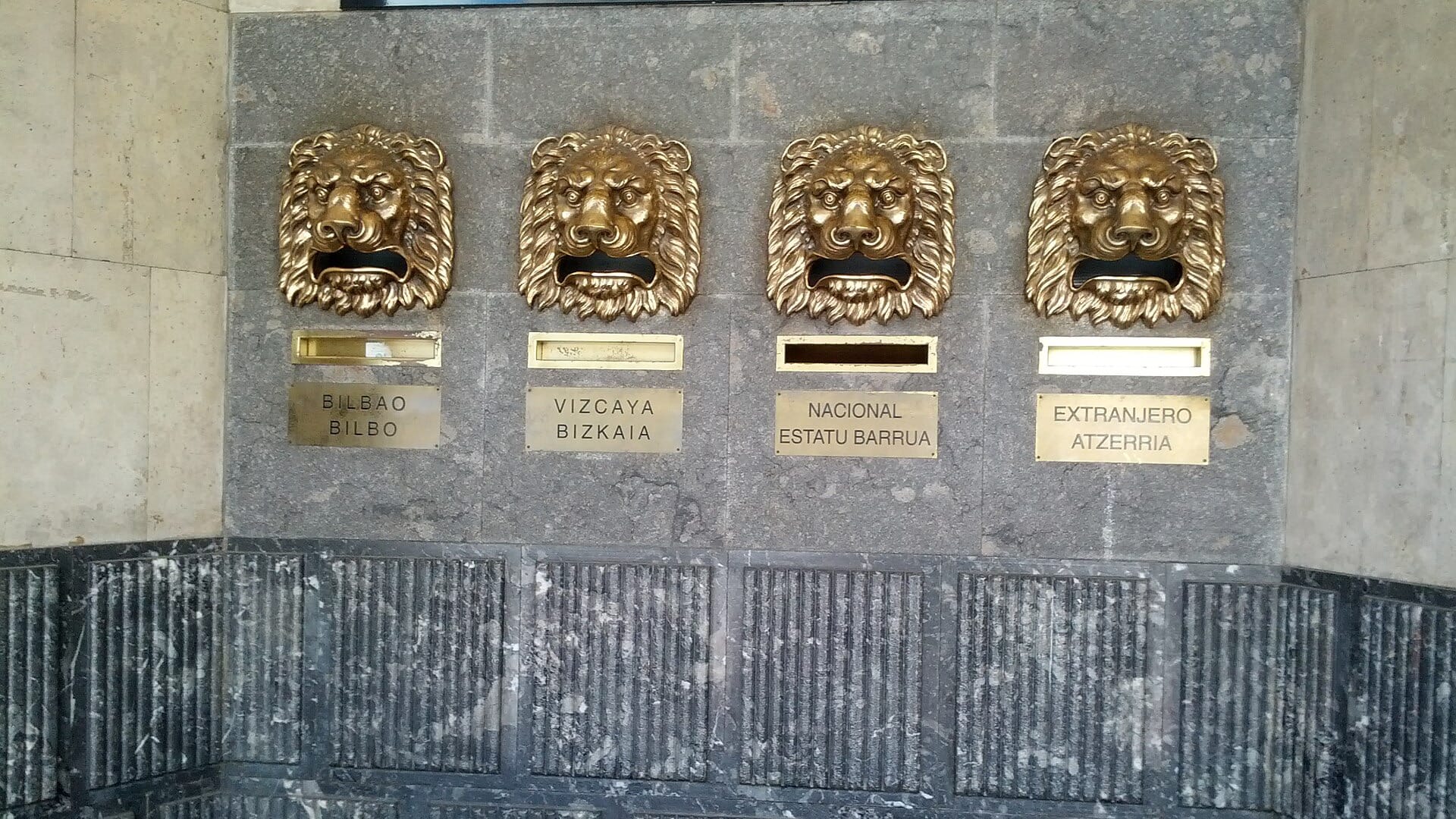 Four brass lion-head mail slots on a stone post-office wall, each labelled with a destination.