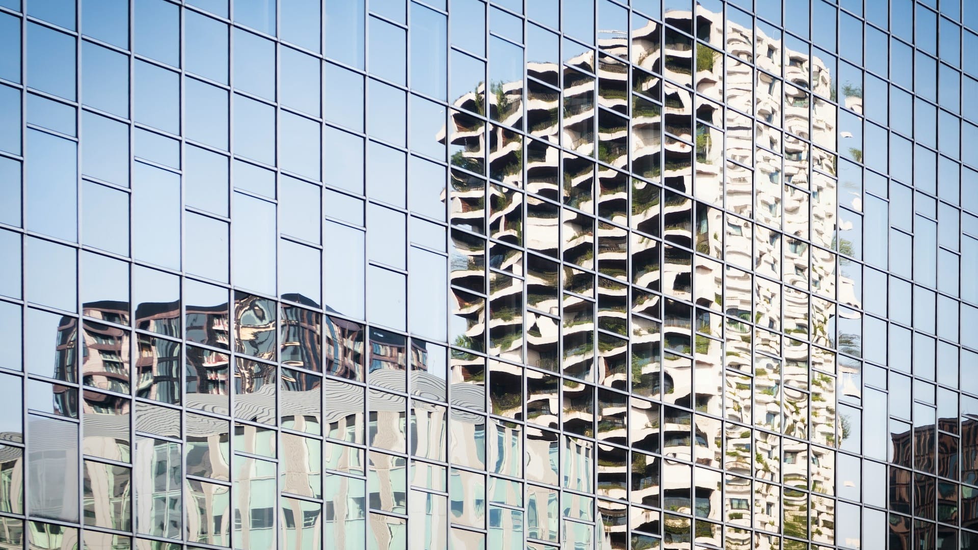 Residential towers reflected in the blue glass grid of an office facade.
