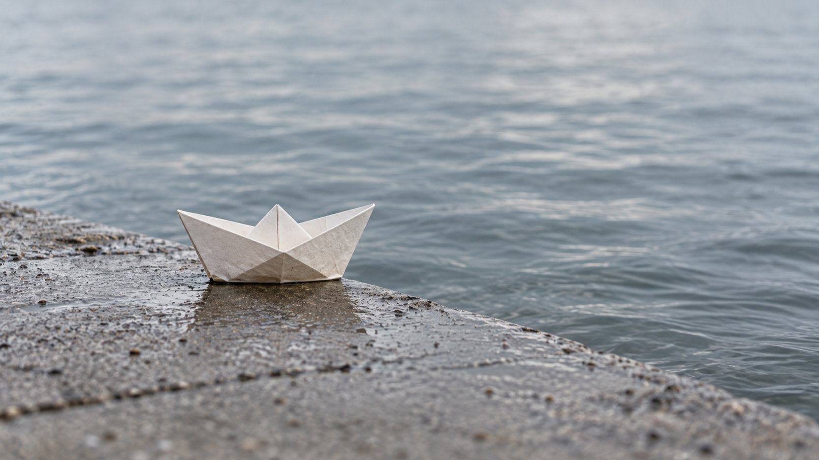 A folded white paper boat waits at the edge of a wet concrete ramp beside calm water.
