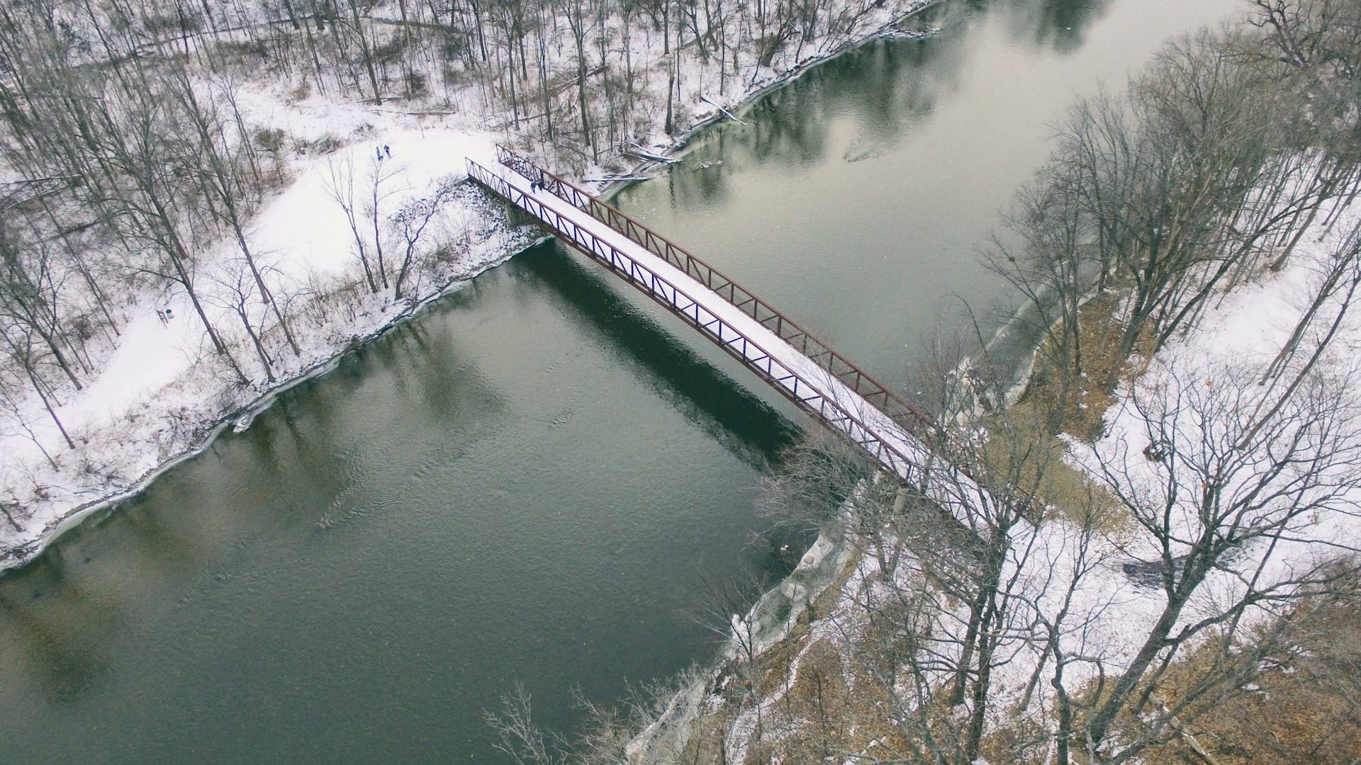 A footbridge crossing a river between snow-covered wooded banks, seen from above.