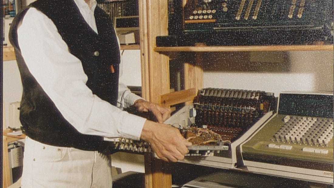 A maintainer repairs an electrical desk computer among shelves of vintage computing equipment.