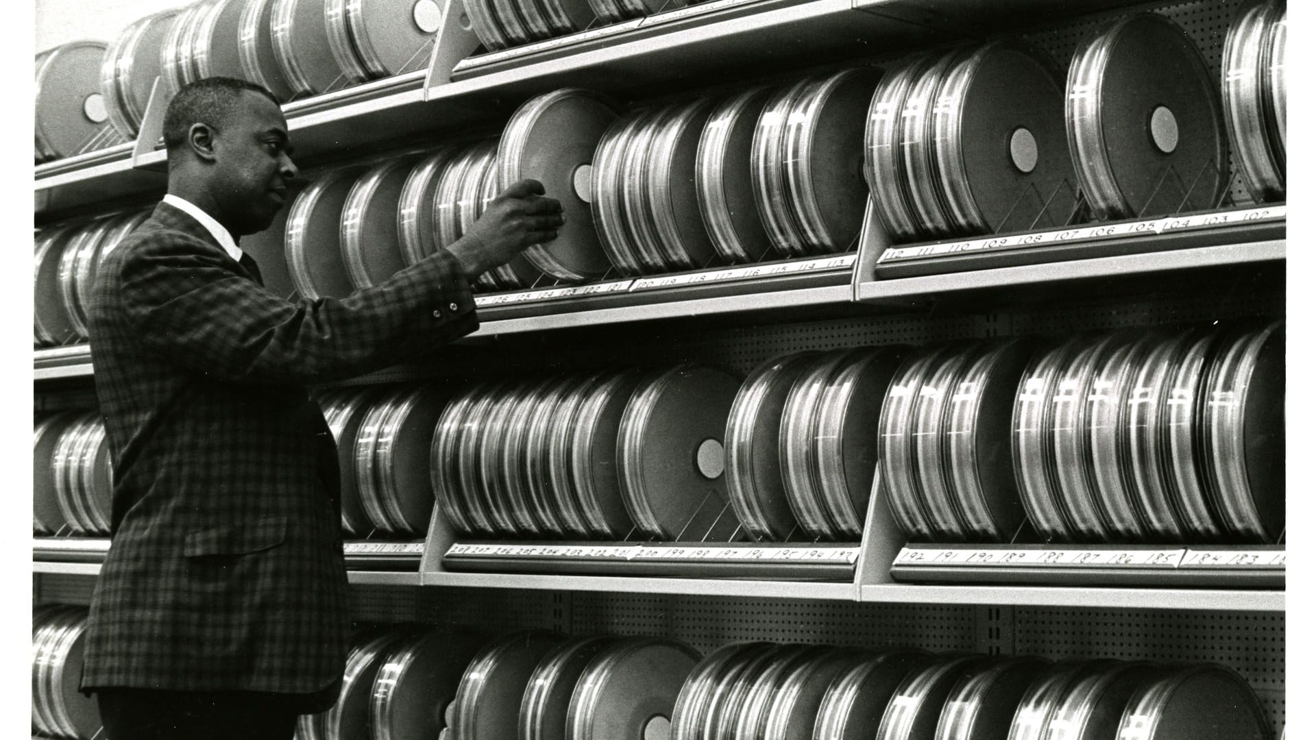 A man in a checked jacket reaches for one of hundreds of magnetic tape reels stacked on shelves in a 1960s computer archive.