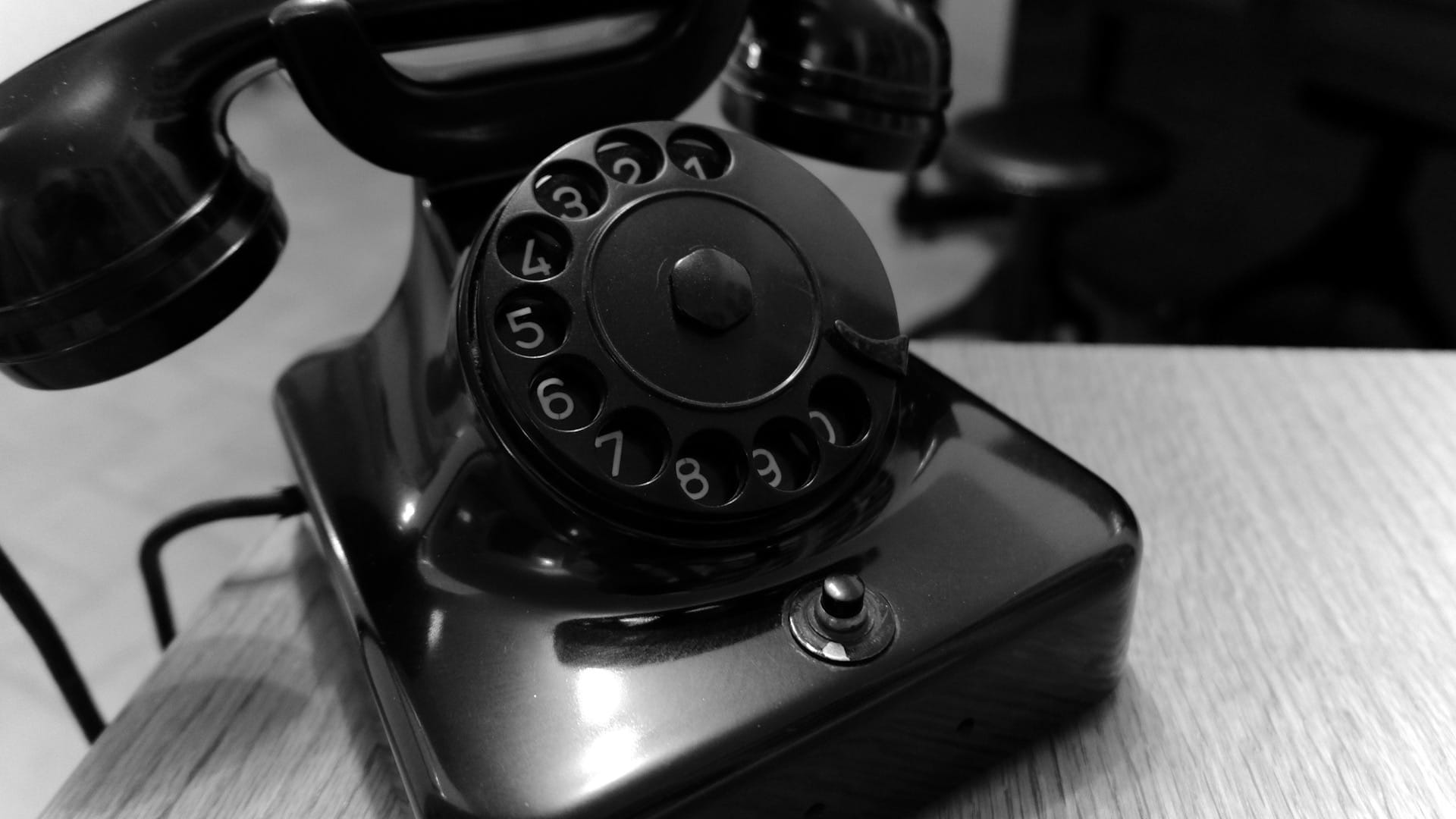 A close-up black-and-white photograph of a rotary telephone on a wooden desk.