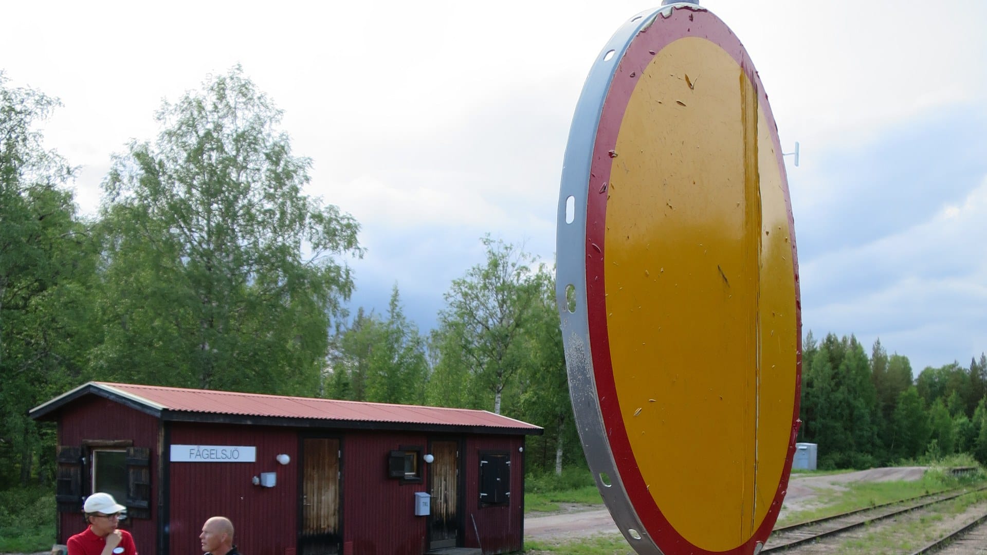 A round yellow-and-red stop-on-request sign beside a small red wooden station building at Fågelsjö, Sweden.