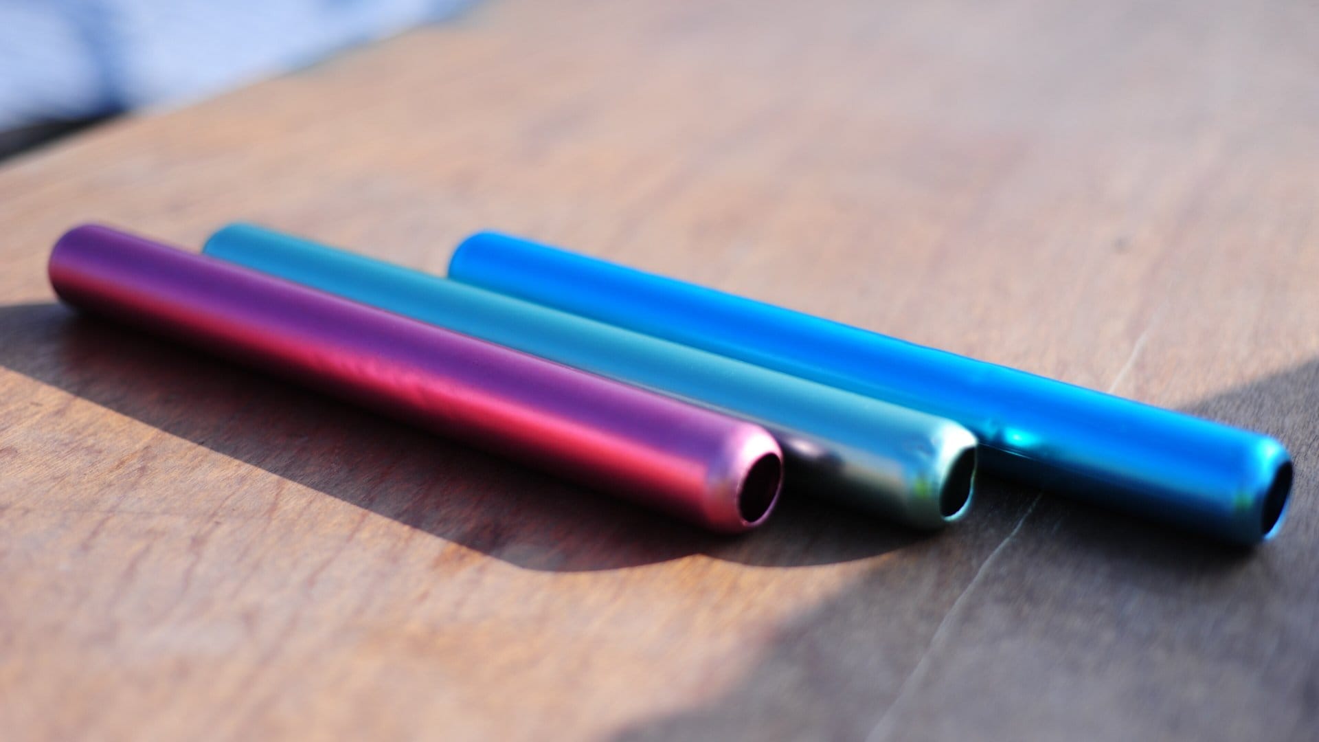 Three aluminium relay batons, purple, teal and blue, lying side by side on a wooden table in low sunlight.