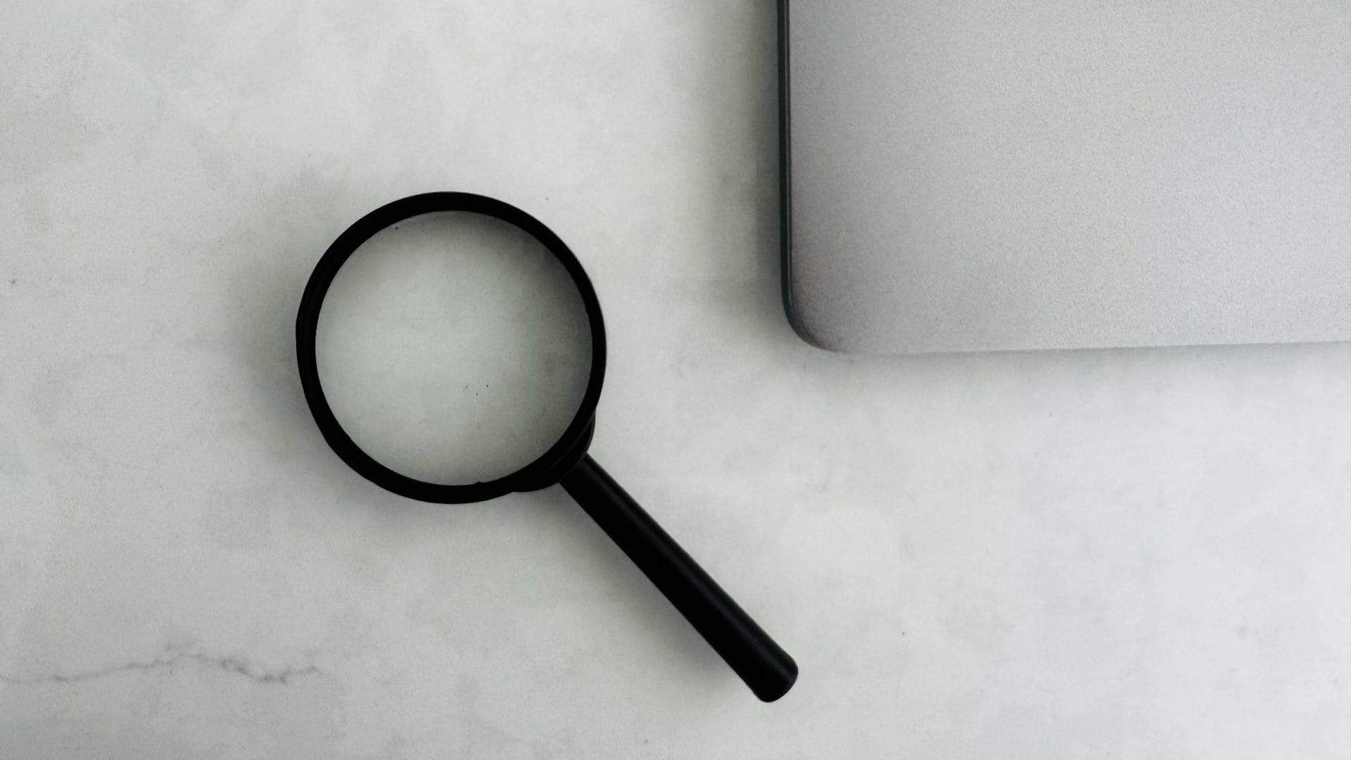 A black magnifying glass beside the corner of a laptop on a pale marble surface.