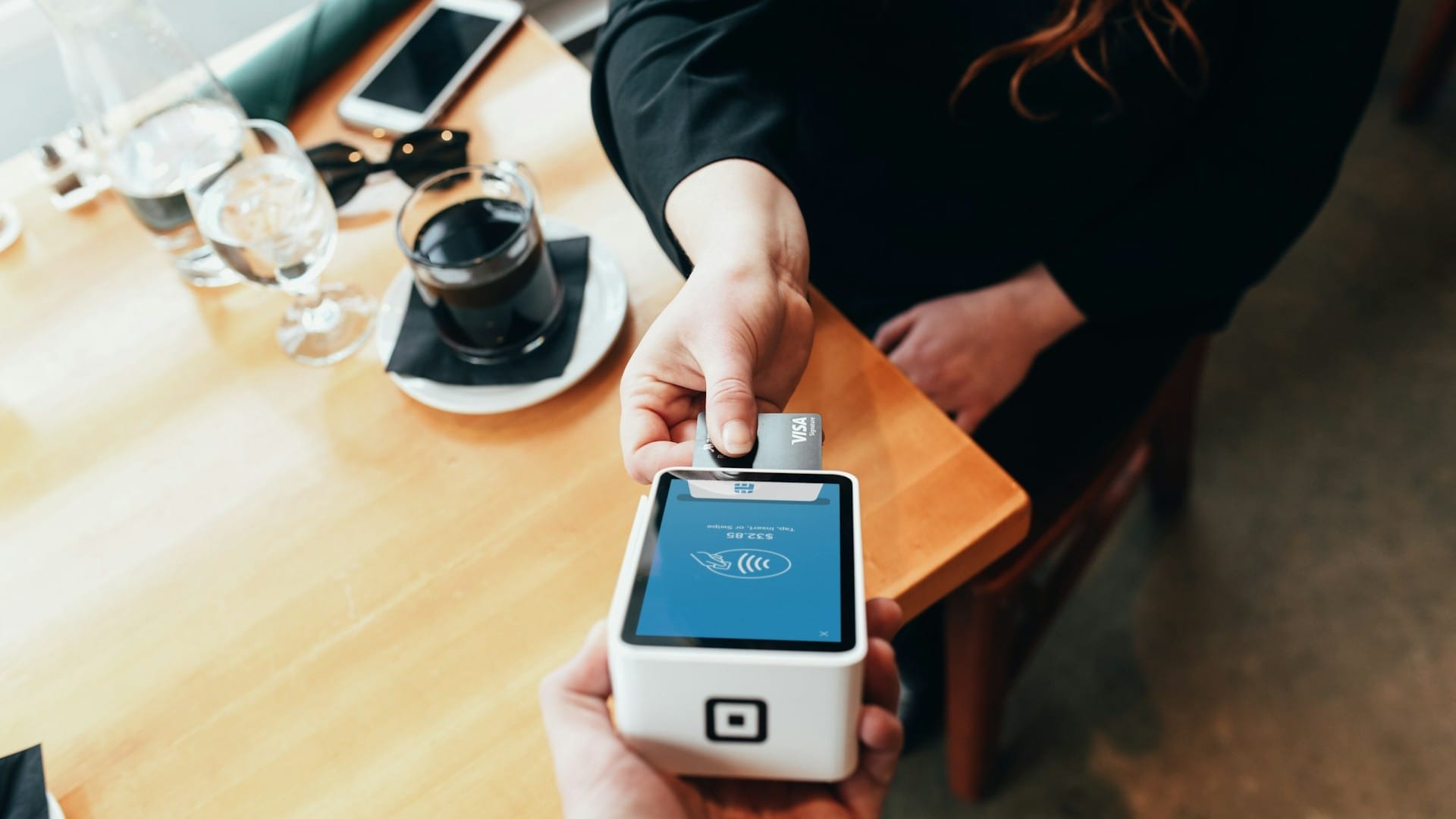 A customer holds a bank card over a handheld payment terminal at a cafe table.