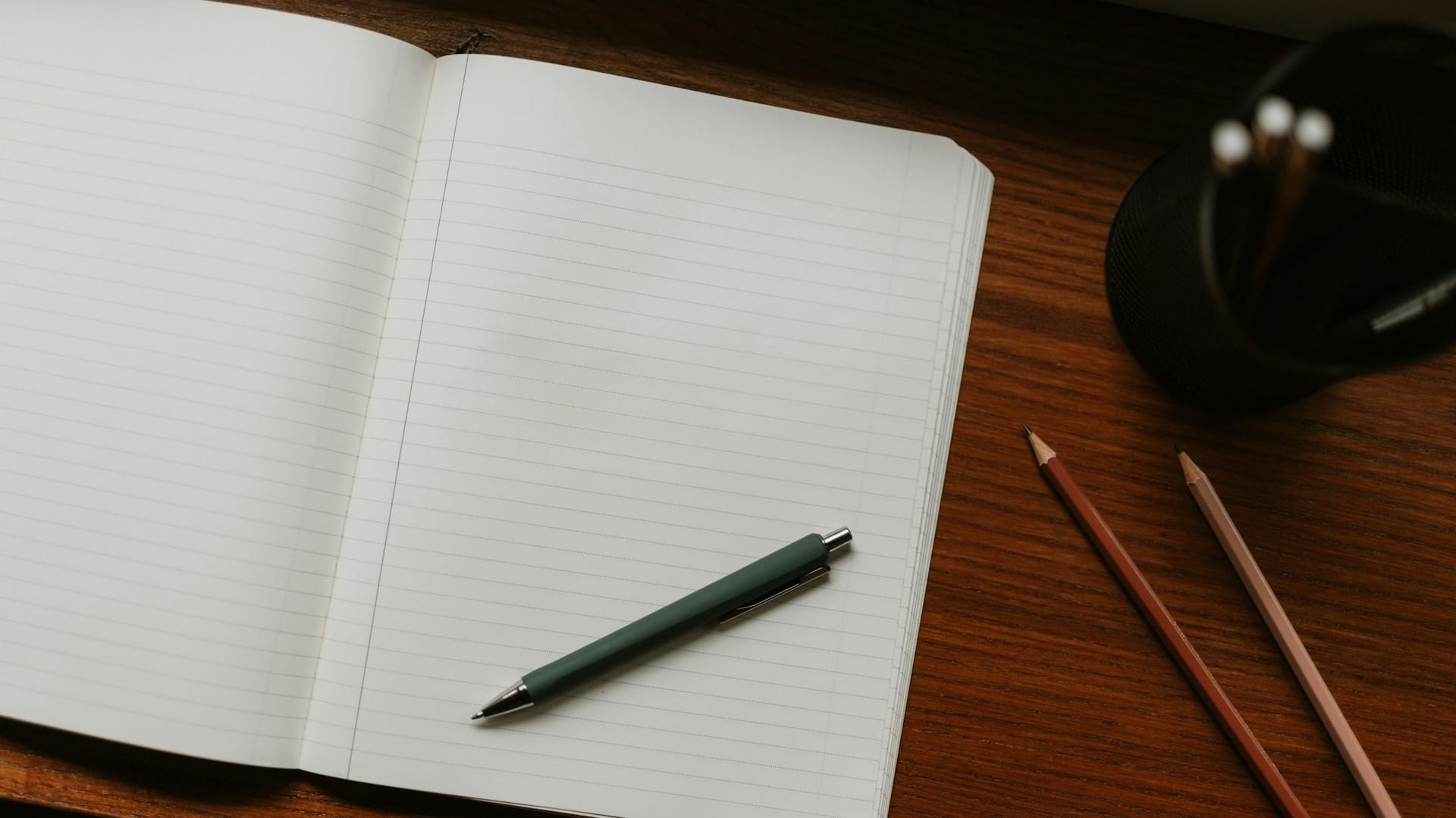 An open lined notebook with a green pen and two pencils on a wooden desk.